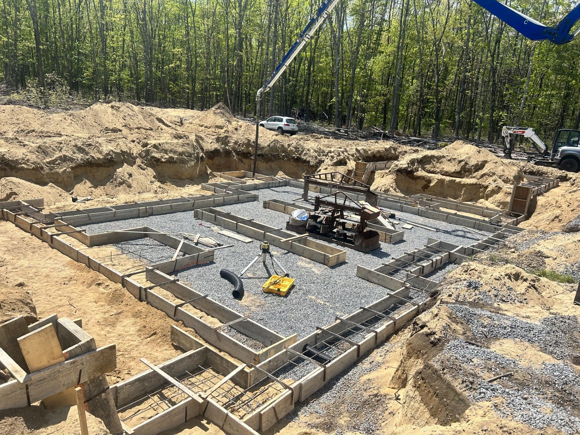 Concrete footings for a building foundation set in a deep, excavated dirt site with a gravel base and woods in the back.
