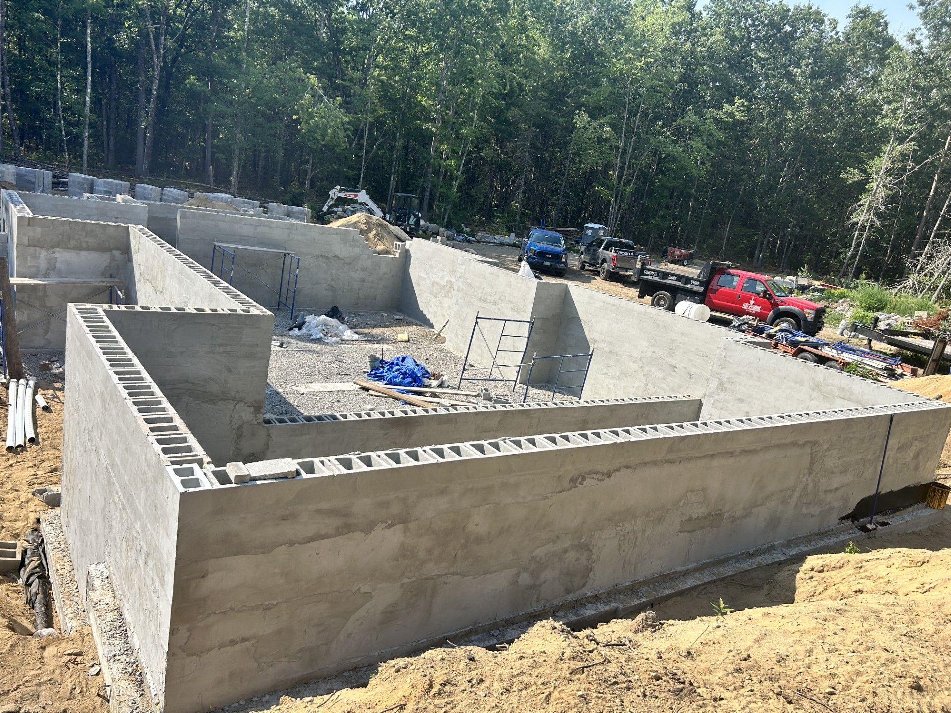 A partially constructed concrete foundation for a new house at a wooded construction site with a red truck in the distance.