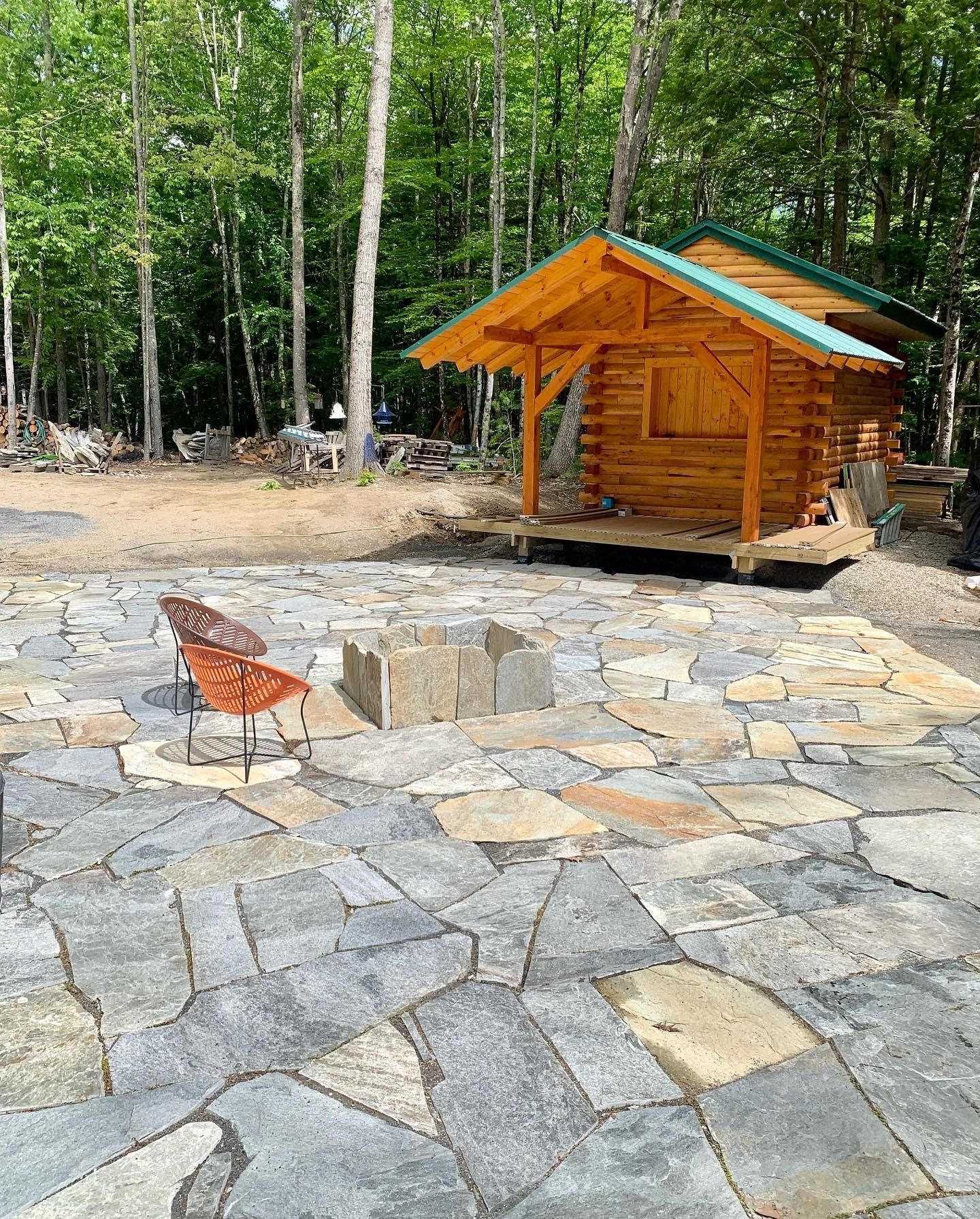 A small wooden log cabin sits in a wooded area behind a flagstone patio with a stone fire pit and two orange chairs.