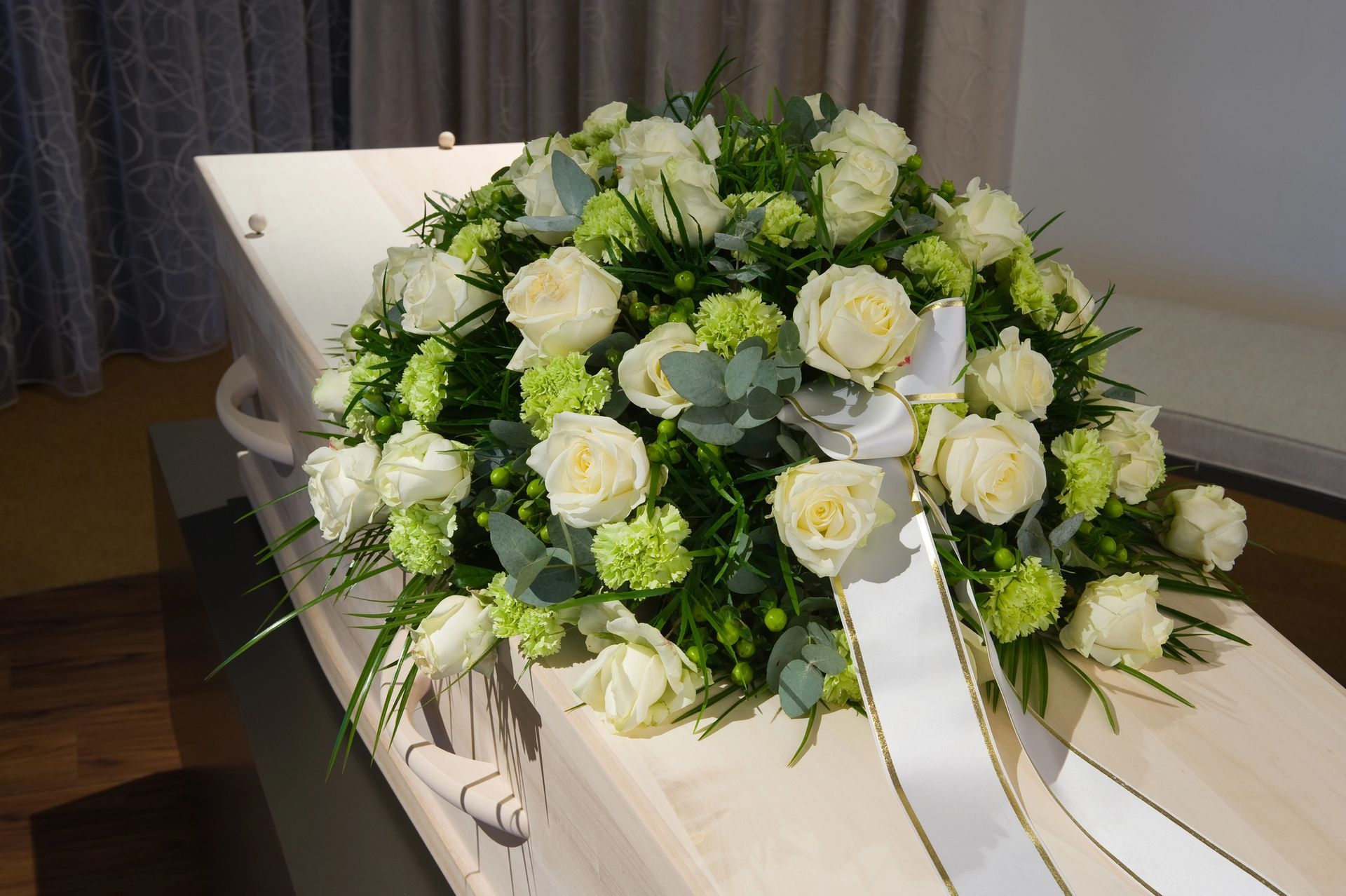 White rose and greenery funeral wreath on a white casket in a dim room