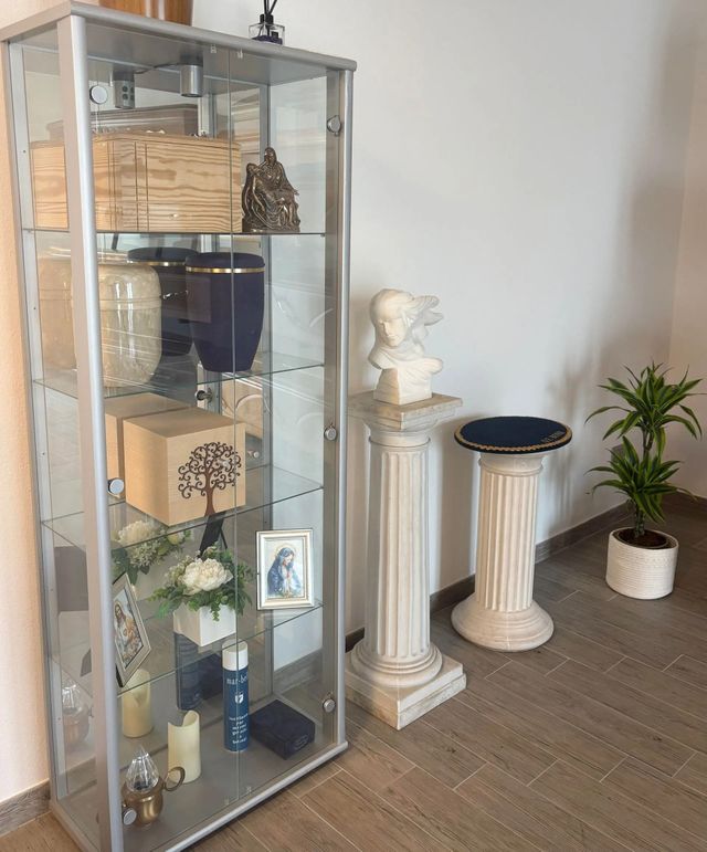Glass display cabinet with decorative boxes, flowers, and candles beside a white pedestal and potted plant