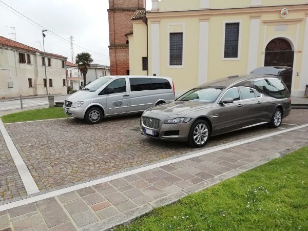 Two parked cars, a silver van and a gray sedan, in a cobblestone courtyard beside a building.