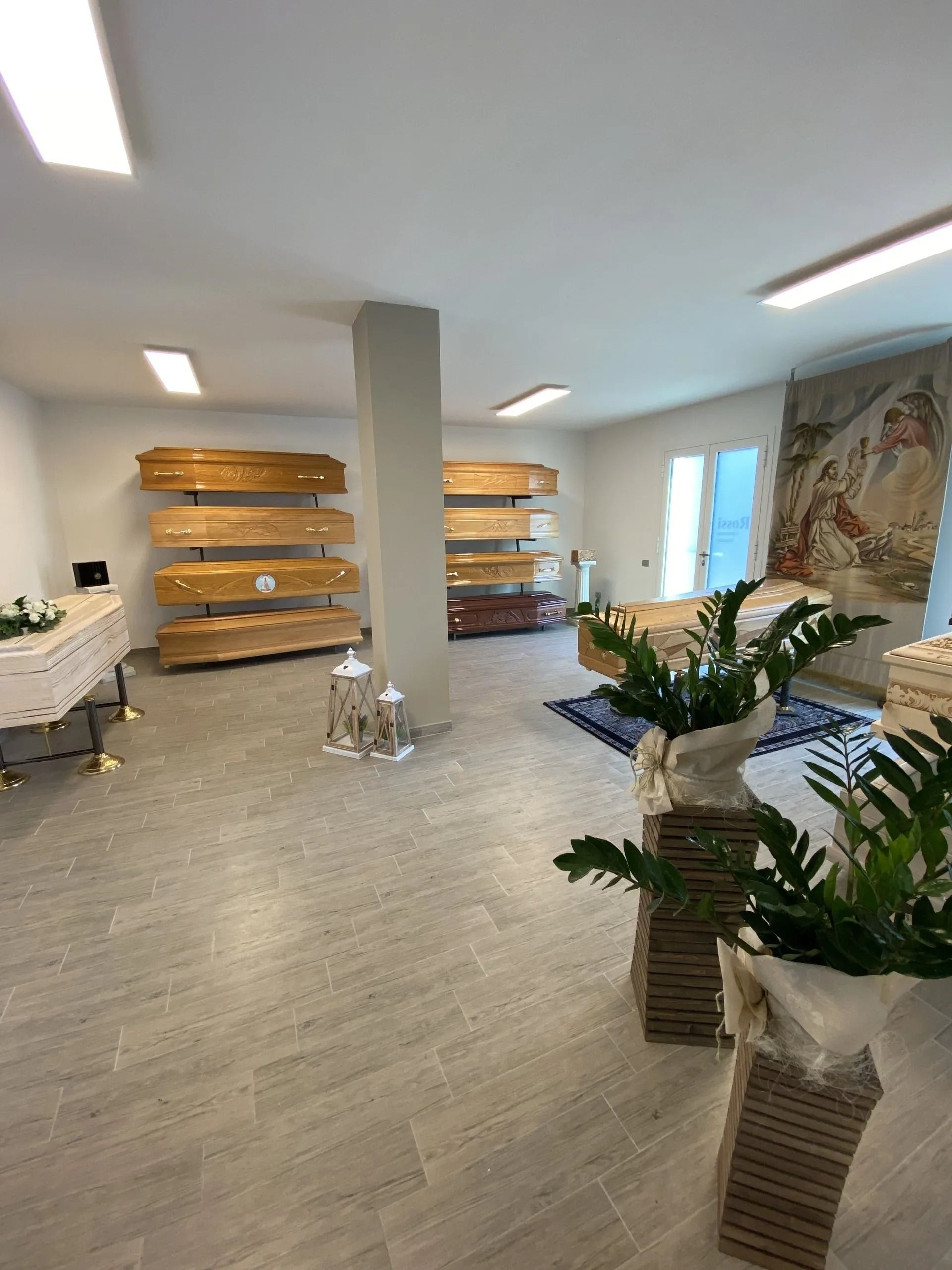 Bright indoor gallery with shelves, potted plants, and a large wall tapestry on a light wood floor