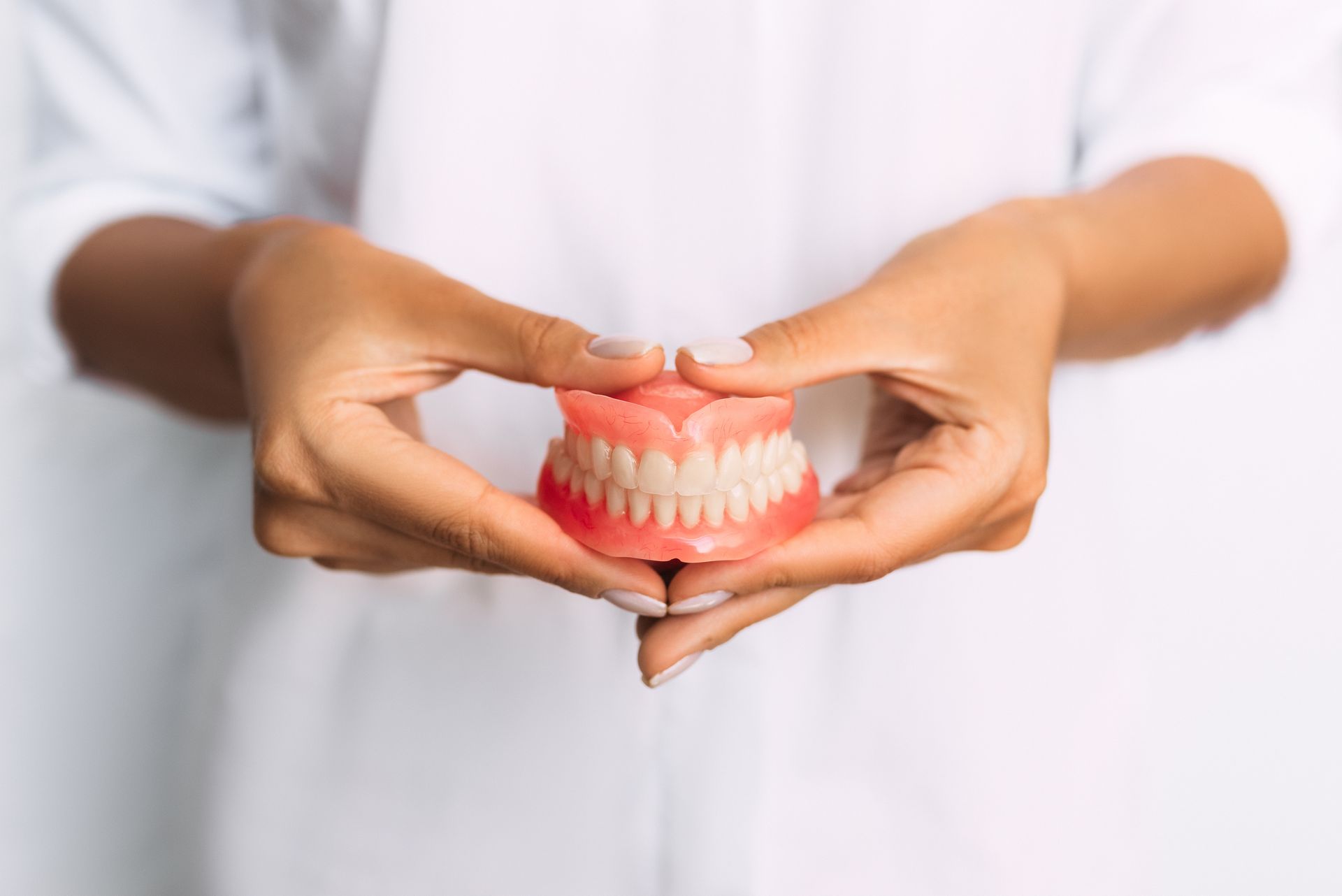 Une personne vêtue d'un haut blanc tient un dentier complet entre ses mains sur un fond blanc uni.