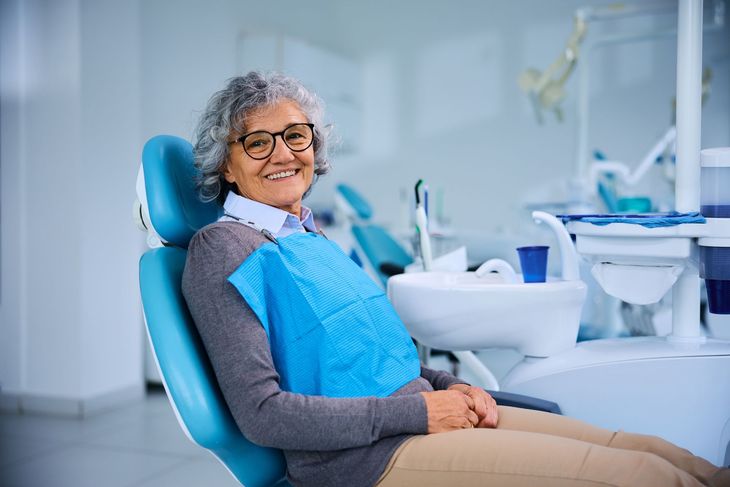 Une personne souriante, portant une bavette dentaire bleue, assise sur un fauteuil dentaire dans une clinique moderne et lumineuse.