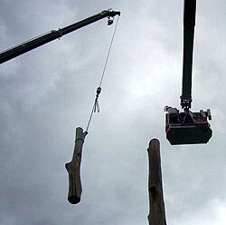 Crane lifts a hanging log between two tall wooden posts against a gray sky