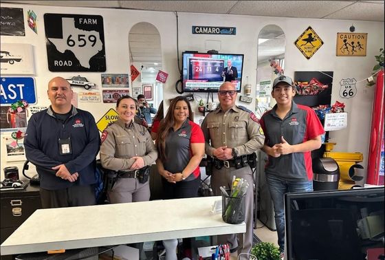 Five people standing in a shop or garage, posing behind a table with tools and signs on the wall.