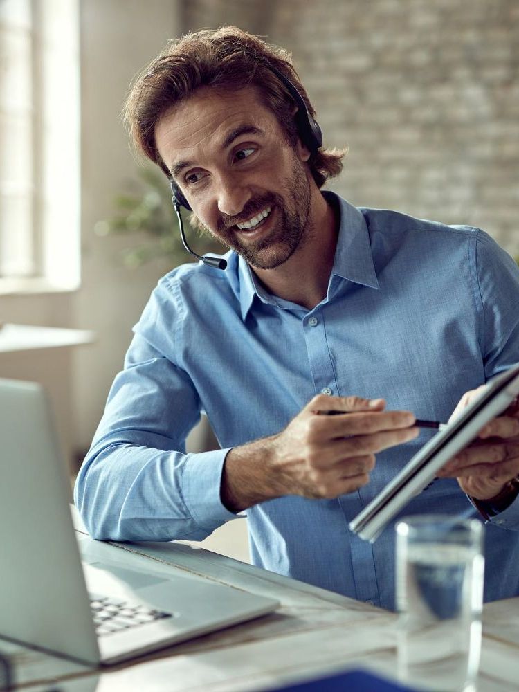 Man in blue shirt on a video call, pointing at notepad, smiling; near laptop, glass of water.