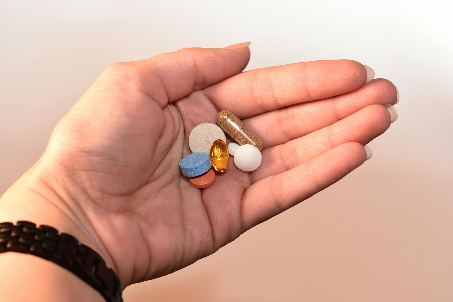 Hand holding a variety of pills, including round and capsule shapes, on a neutral background.