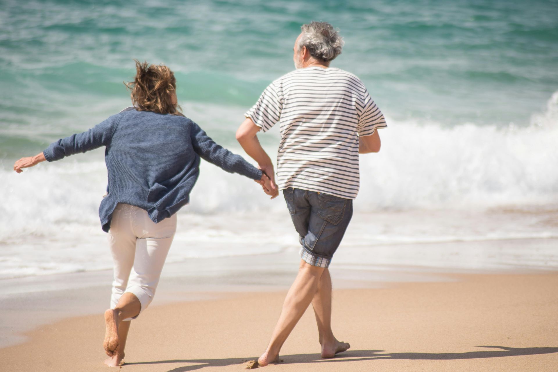 Elderly couple running hand-in-hand on a beach, near ocean waves.