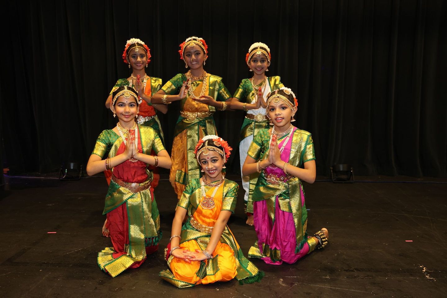 Six Kuchipudi performers in colorful traditional costumes pose on a stage against a dark curtain.
