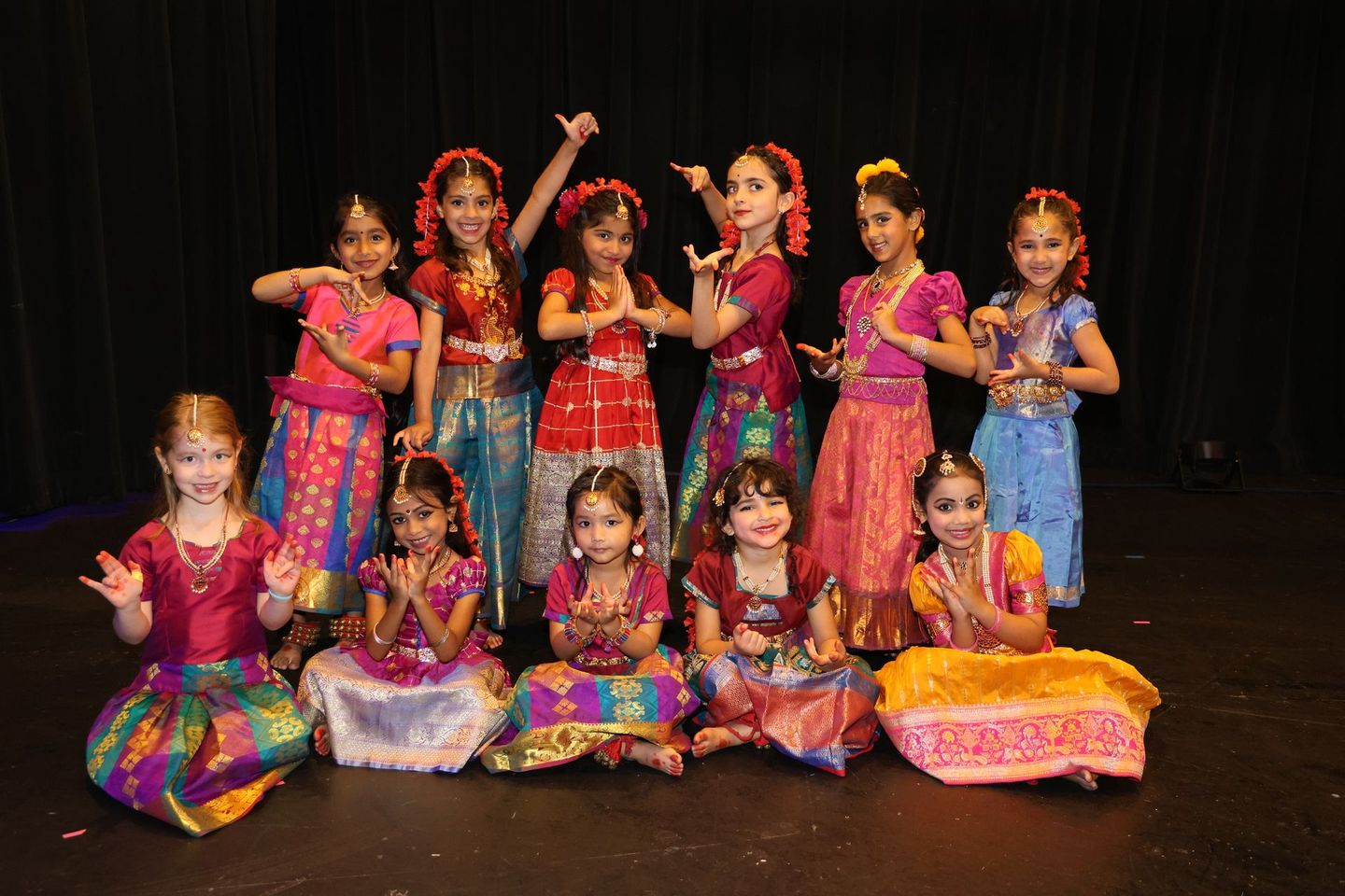 Group of children in colorful traditional dance costumes posing on a stage with a dark backdrop