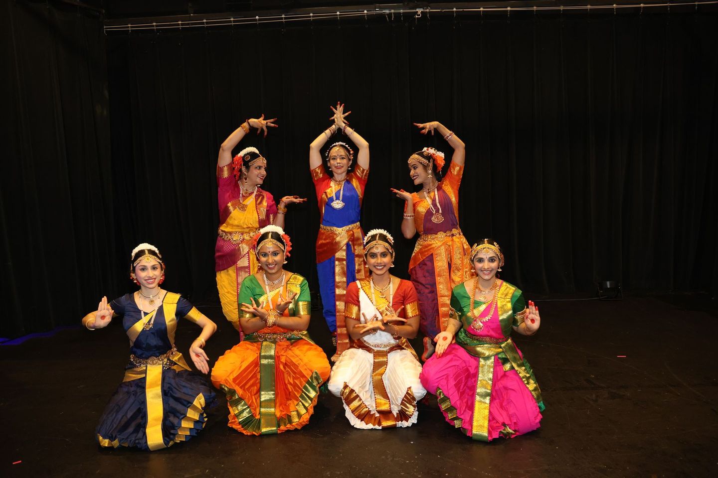 Seven Kuchipudi dancers in colorful traditional costumes pose on a dark stage, three standing and four seated.