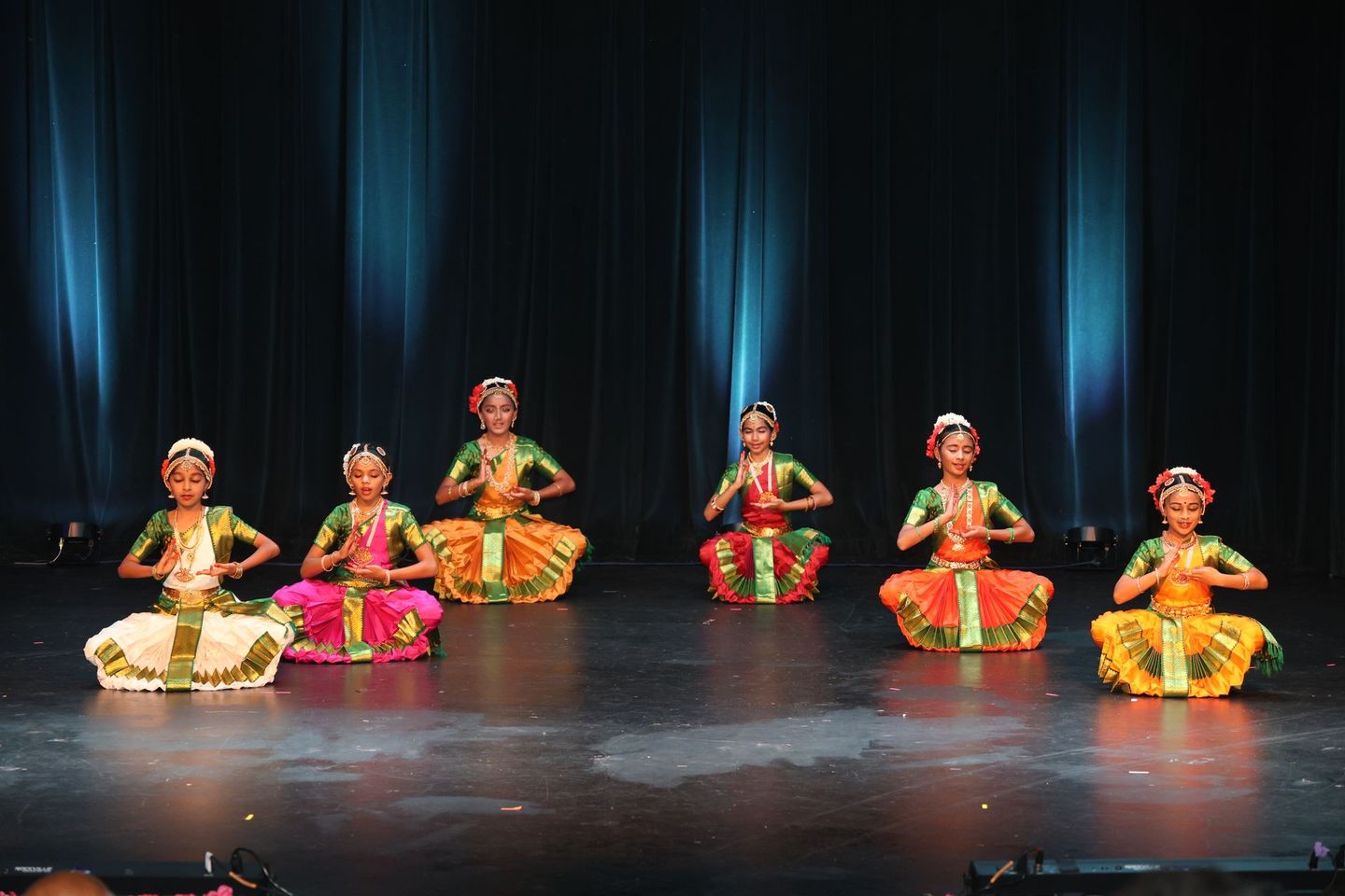 Six seated Kuchipudi dancers in bright costumes performing on a dark stage with blue lights