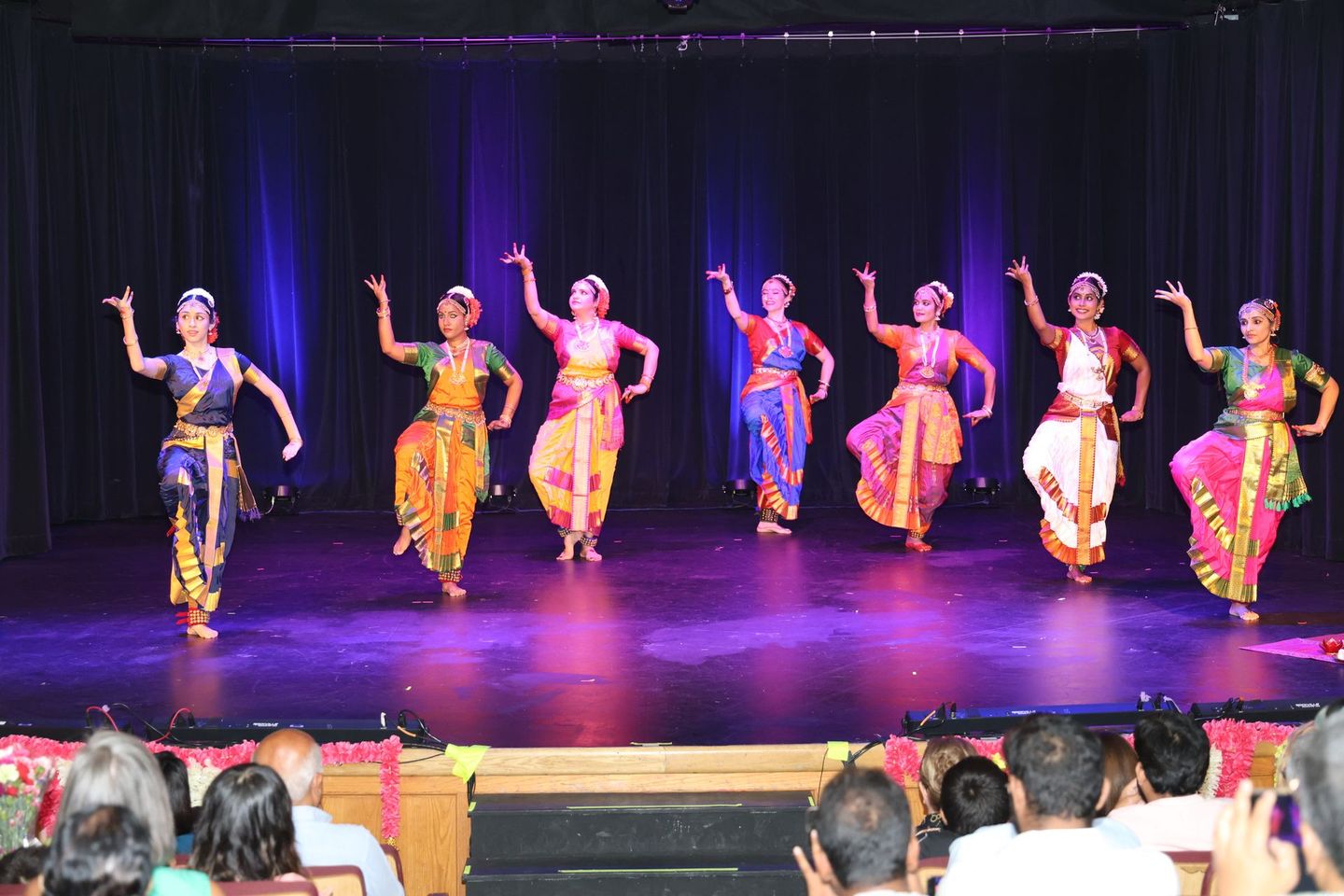 Seven Kuchipudi dancers in colorful costumes performing on a stage before an audience