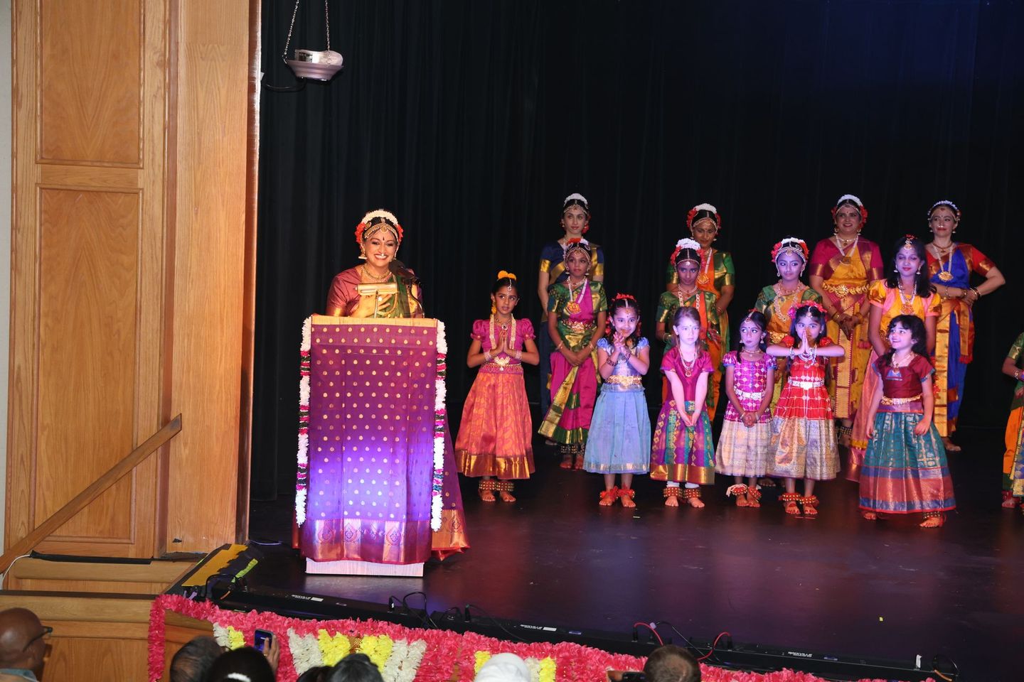 Children in colorful costumes on a theater stage, with one person at a podium and floral decorations in front.