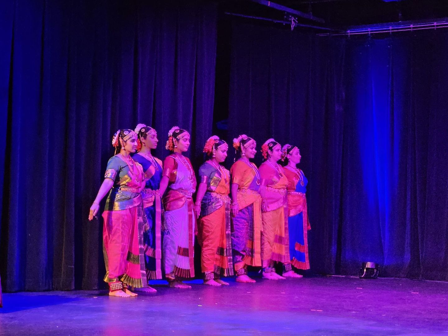 A group of Kuchipudi dancers in colorful traditional outfits performing on a stage with purple lighting and black curtains