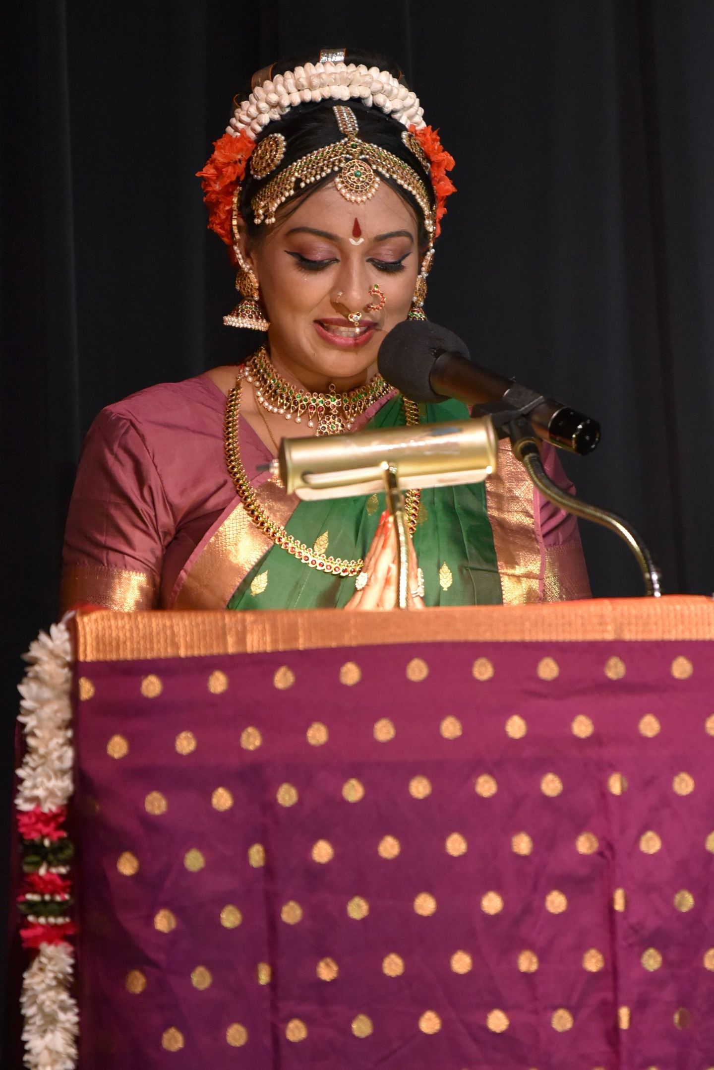 Kuchipudi dancer in green sari speaking at a podium onstage, wearing gold jewelry and flowers in her hair.