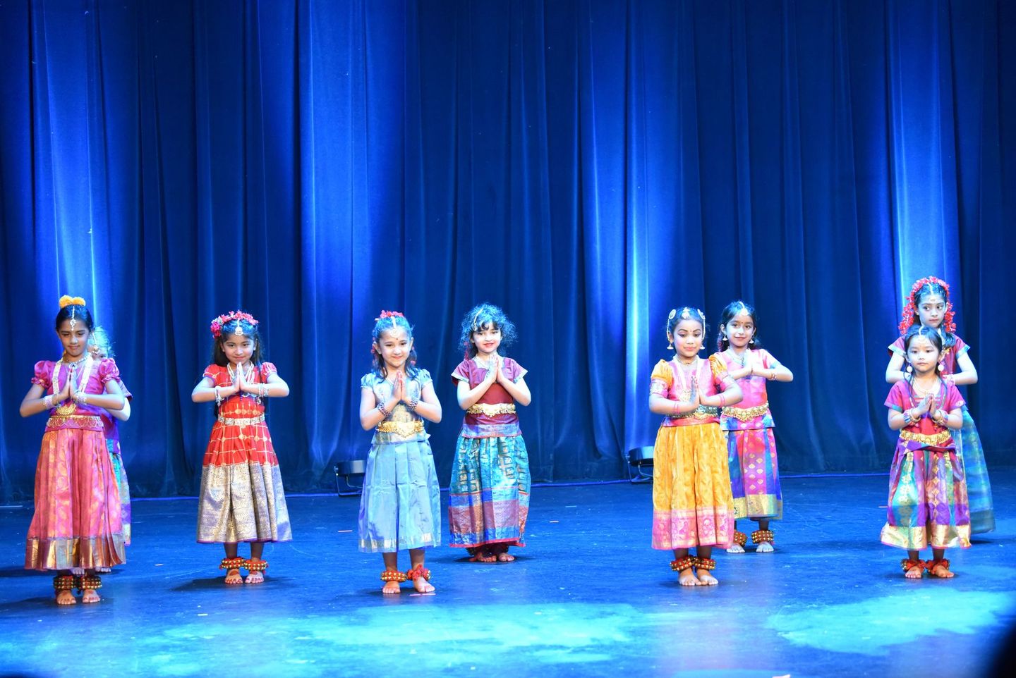 Seven children in colorful costumes stand on a stage with blue curtains, hands together in a dance pose.