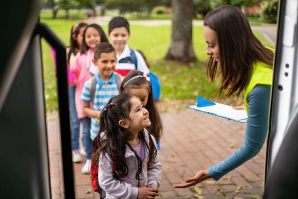 Children line up to board a bus, assisted by an adult in a high-visibility vest. The setting appears to be a school or park.