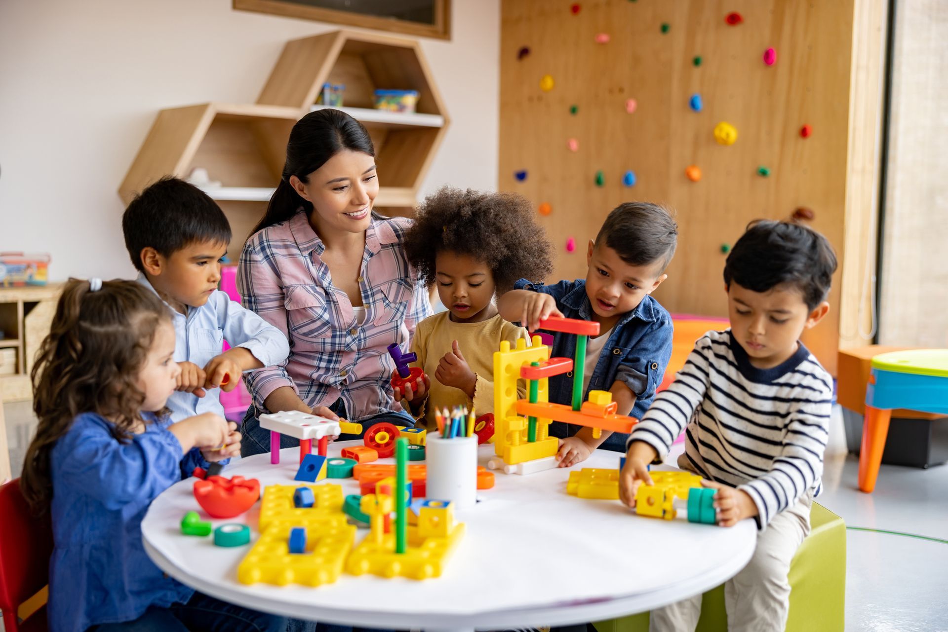 A teacher and six children play with colorful blocks around a white table in a brightly lit classroom.