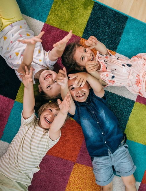 Four young children smiling and reaching upwards while lying together on a colorful patchwork carpet.
