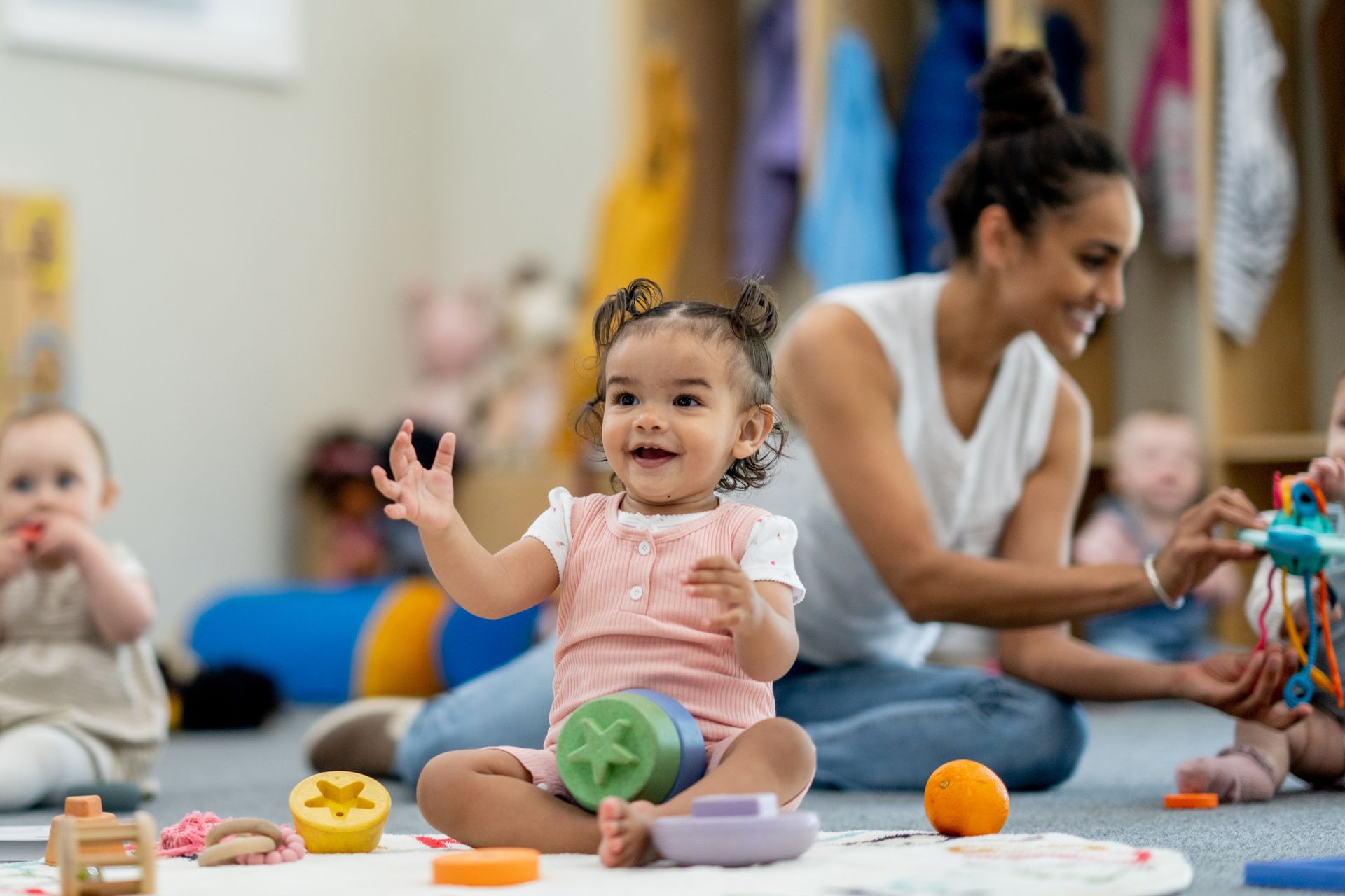 A young child smiles, reaching for a toy on a play mat with a caregiver in a daycare setting. Other babies and toys are nearby.