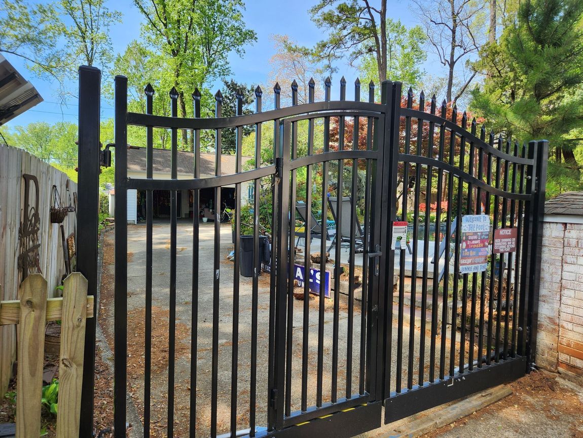 Black metal gate at a gated driveway entrance with trees and a house in the background