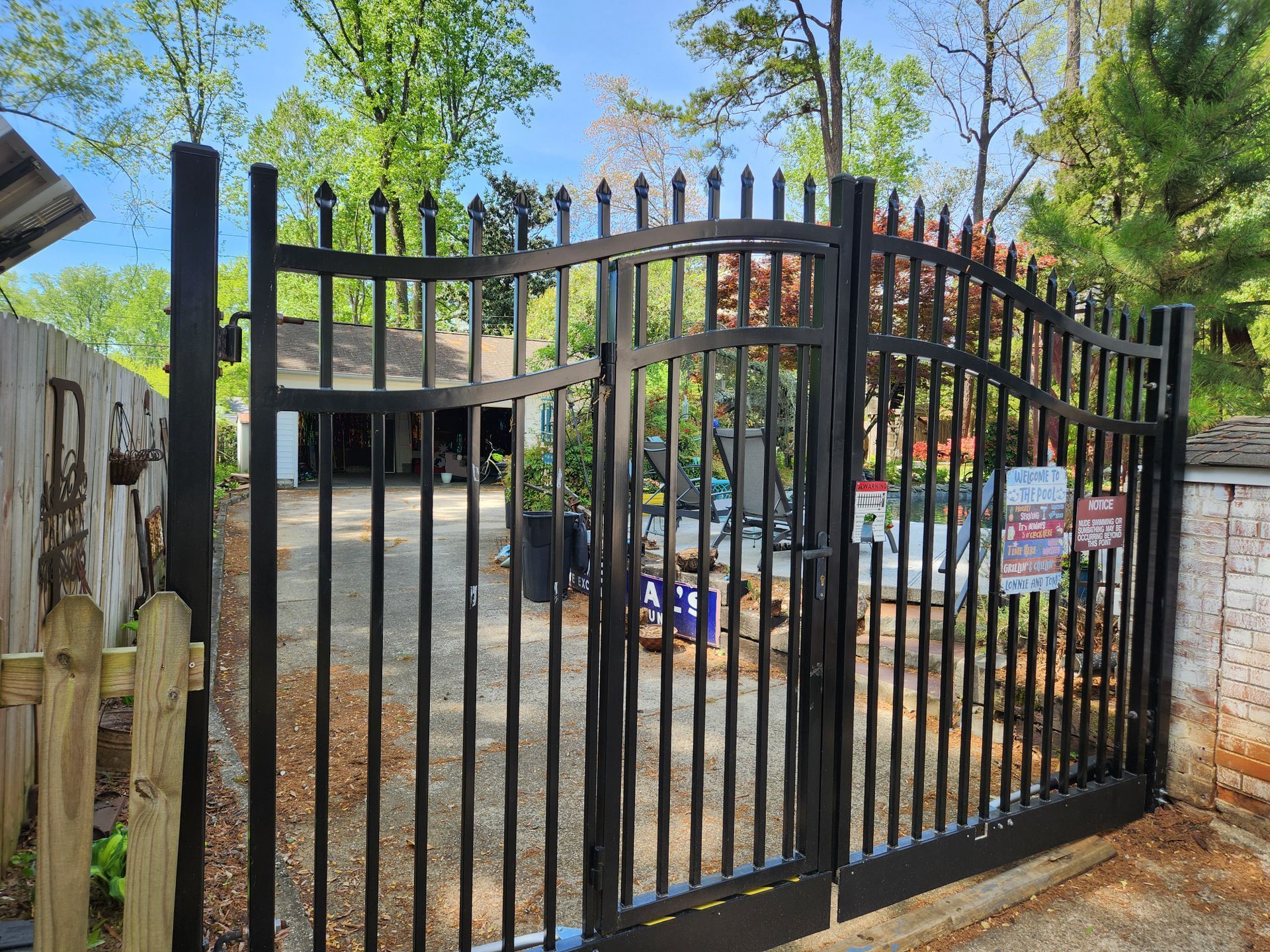 Black metal gate at a fenced entrance to a yard with trees and a house in the background