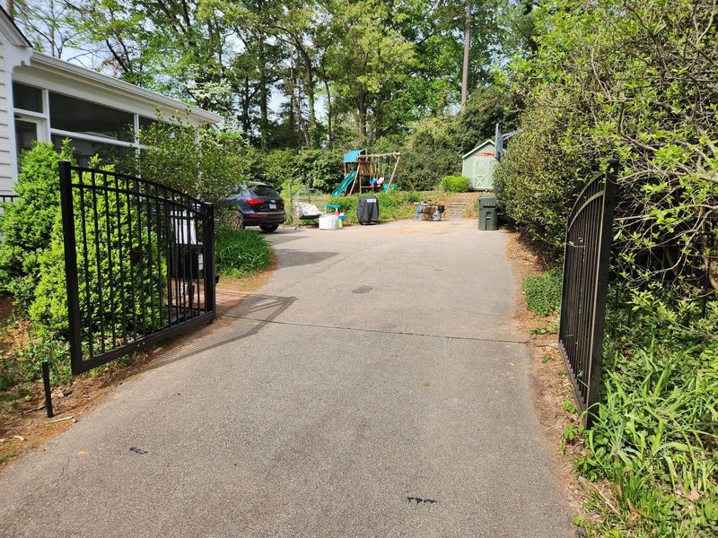 Gravel driveway with open black gates leading to a house and trees in a wooded yard