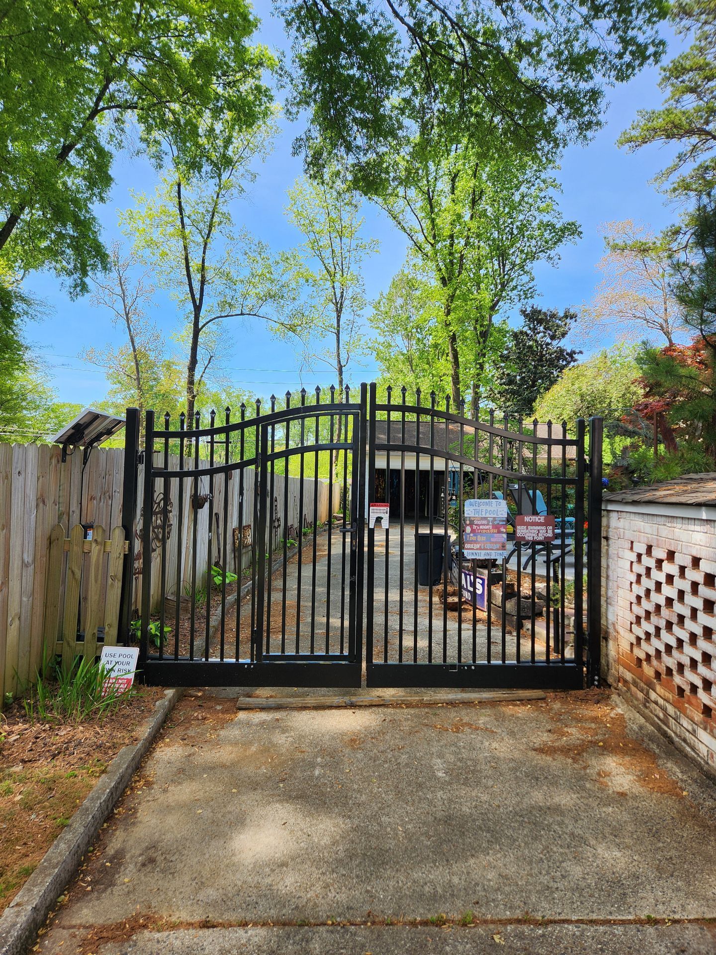 Black metal gate at the end of a concrete driveway with trees overhead and yard items beyond it