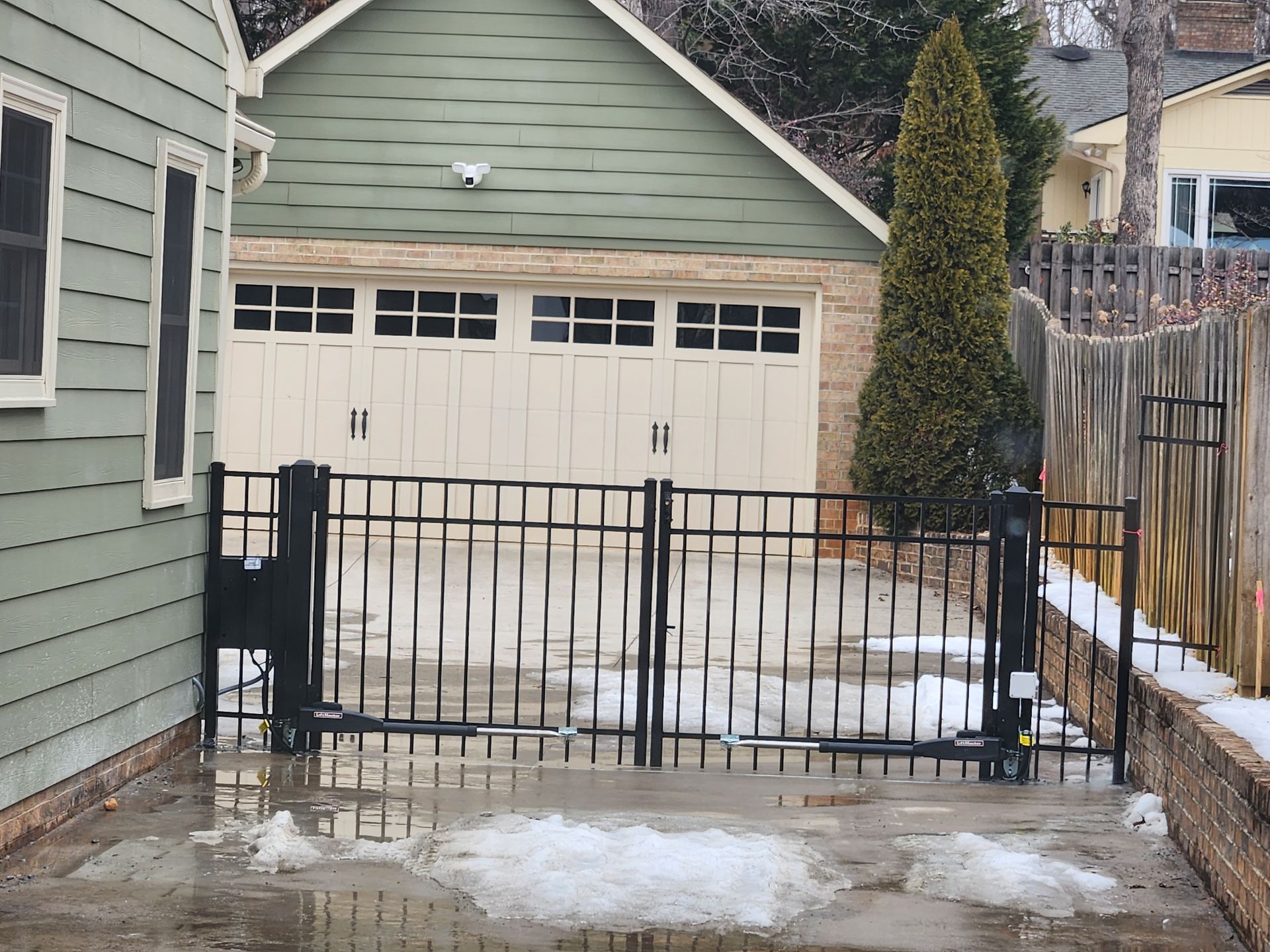 Snowy driveway with a black metal gate in front of a garage and green house siding