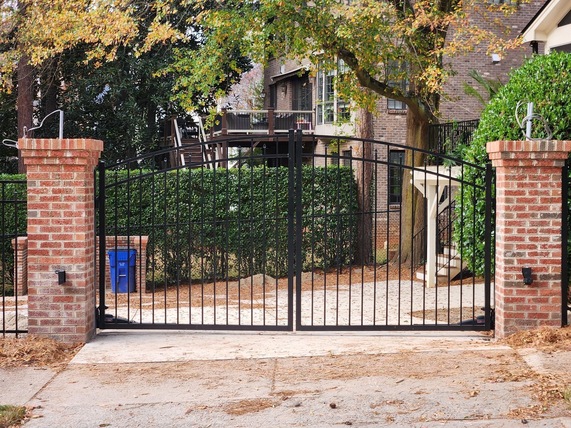 Black wrought-iron gate between two brick pillars at a driveway entrance under leafy trees.