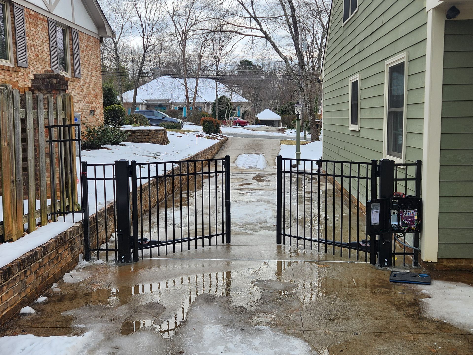 Black metal gate across a wet driveway between houses in a snowy yard