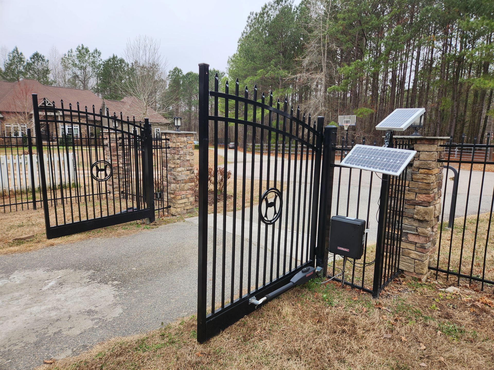 Open black wrought-iron gate by a gravel driveway with stone pillars and solar panels