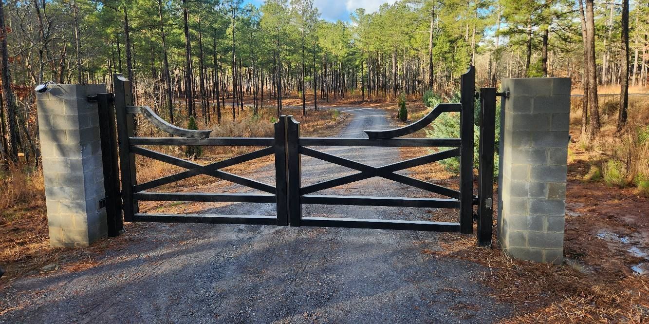 Black wooden gate across a gravel driveway between stone pillars in a wooded area