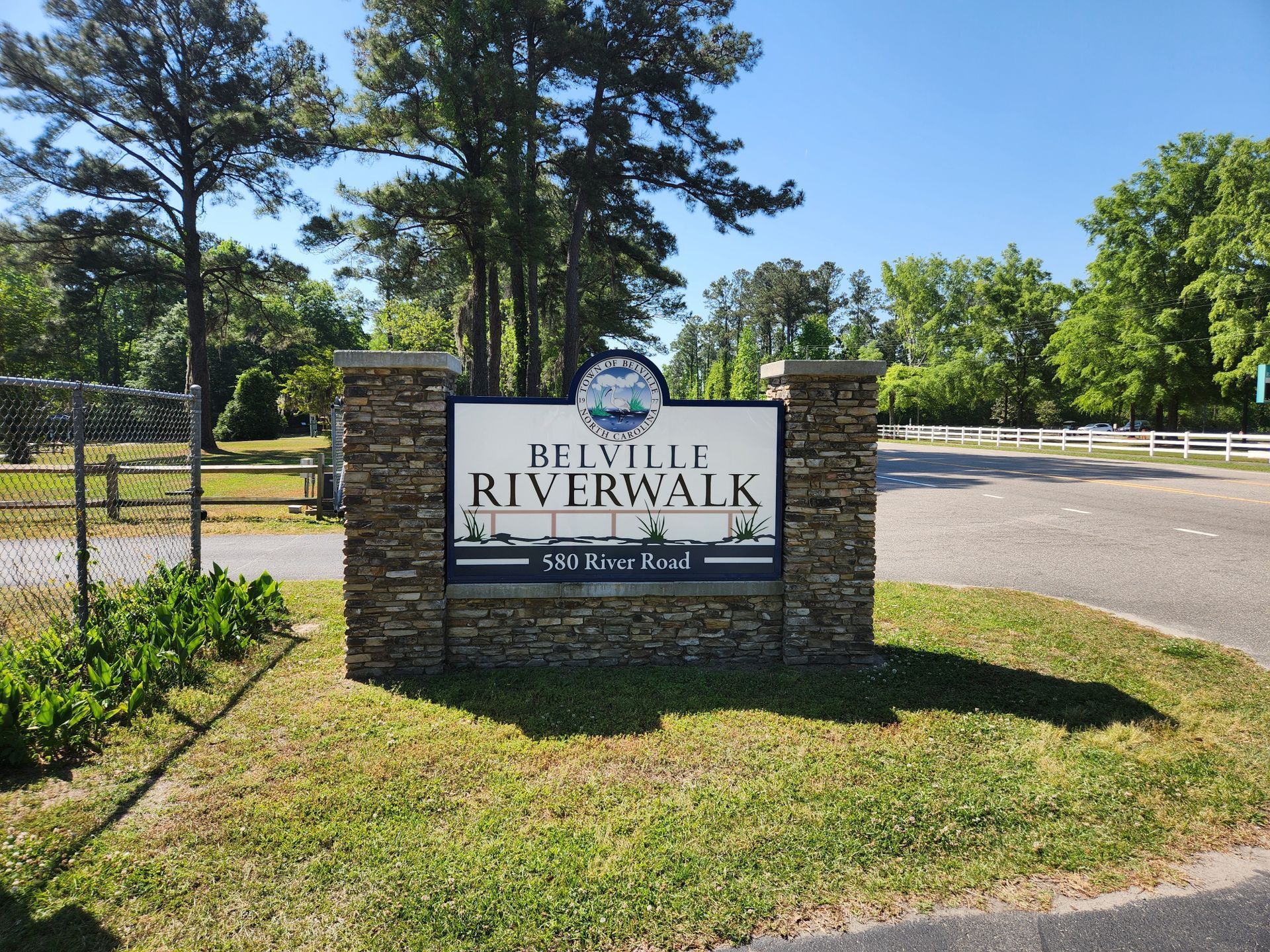 Stone sign reading “Riverwalk” beside a road, with grass, trees, and a clear blue sky.