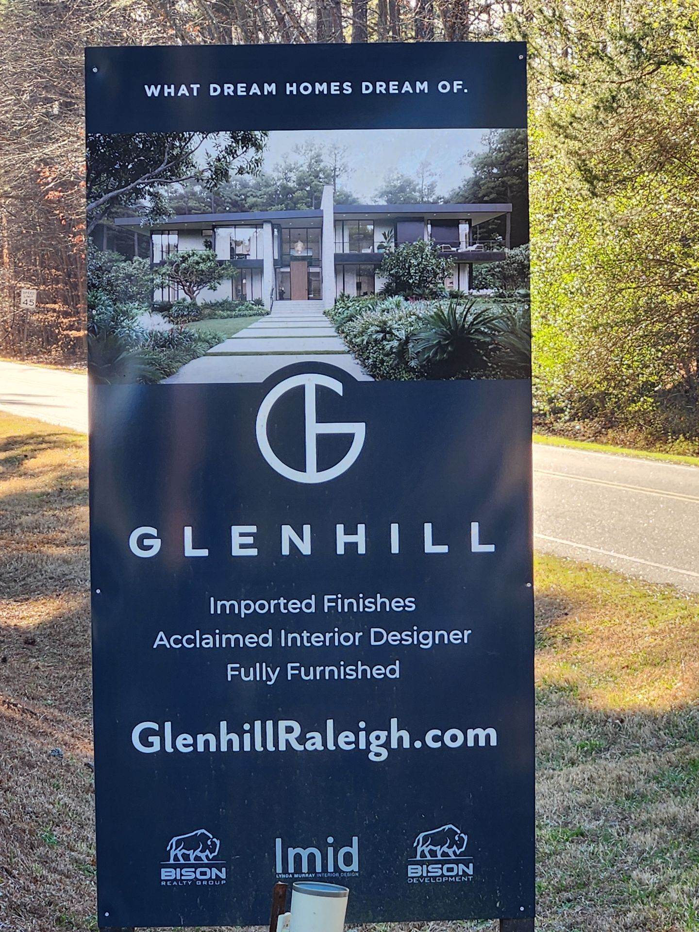 Dark blue Glenhill real estate sign with home photo and website beside a tree-lined road.