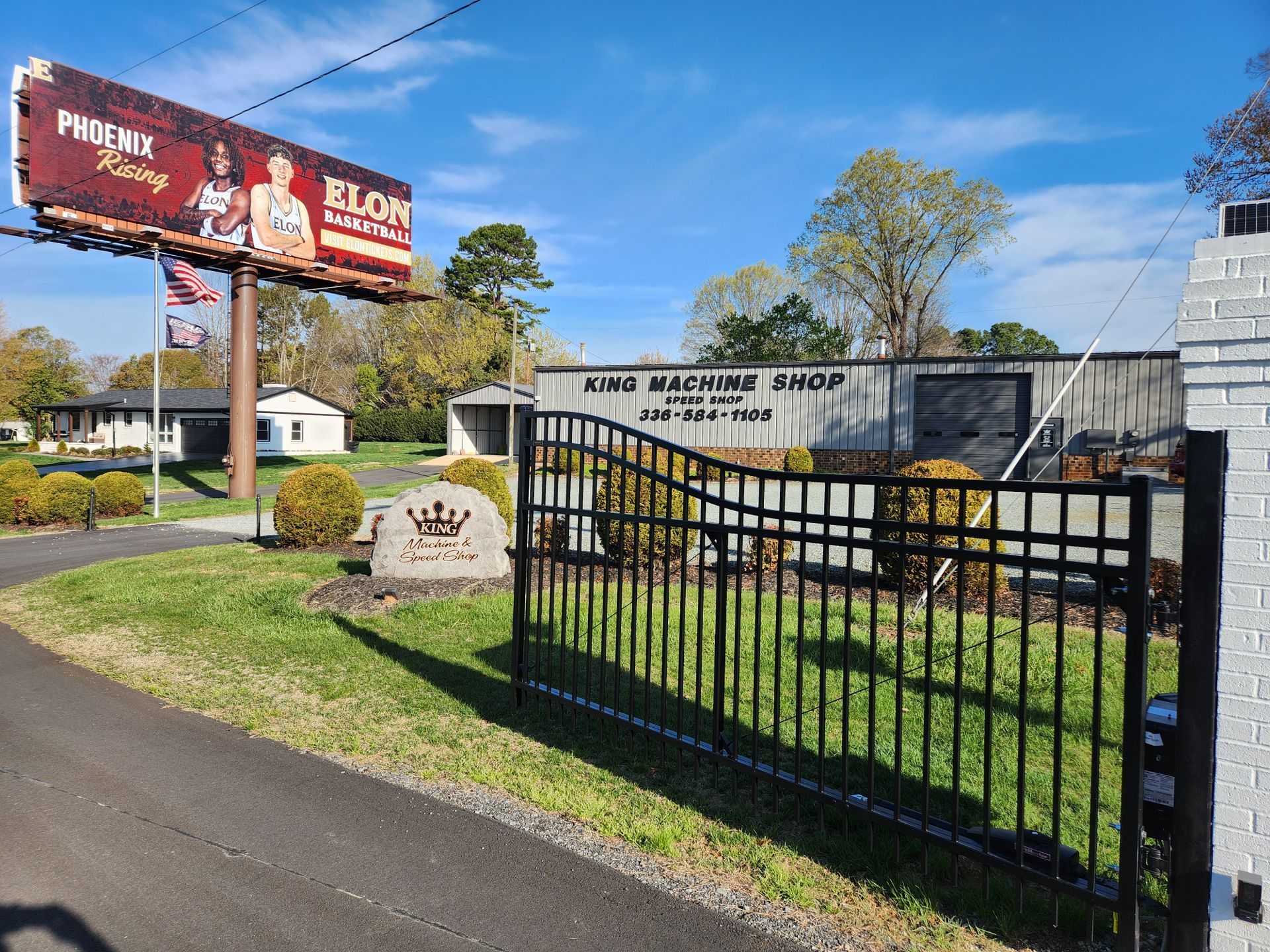 Black metal gate at a roadside business with sign reading “King Machine Shop,” near a red billboard and house