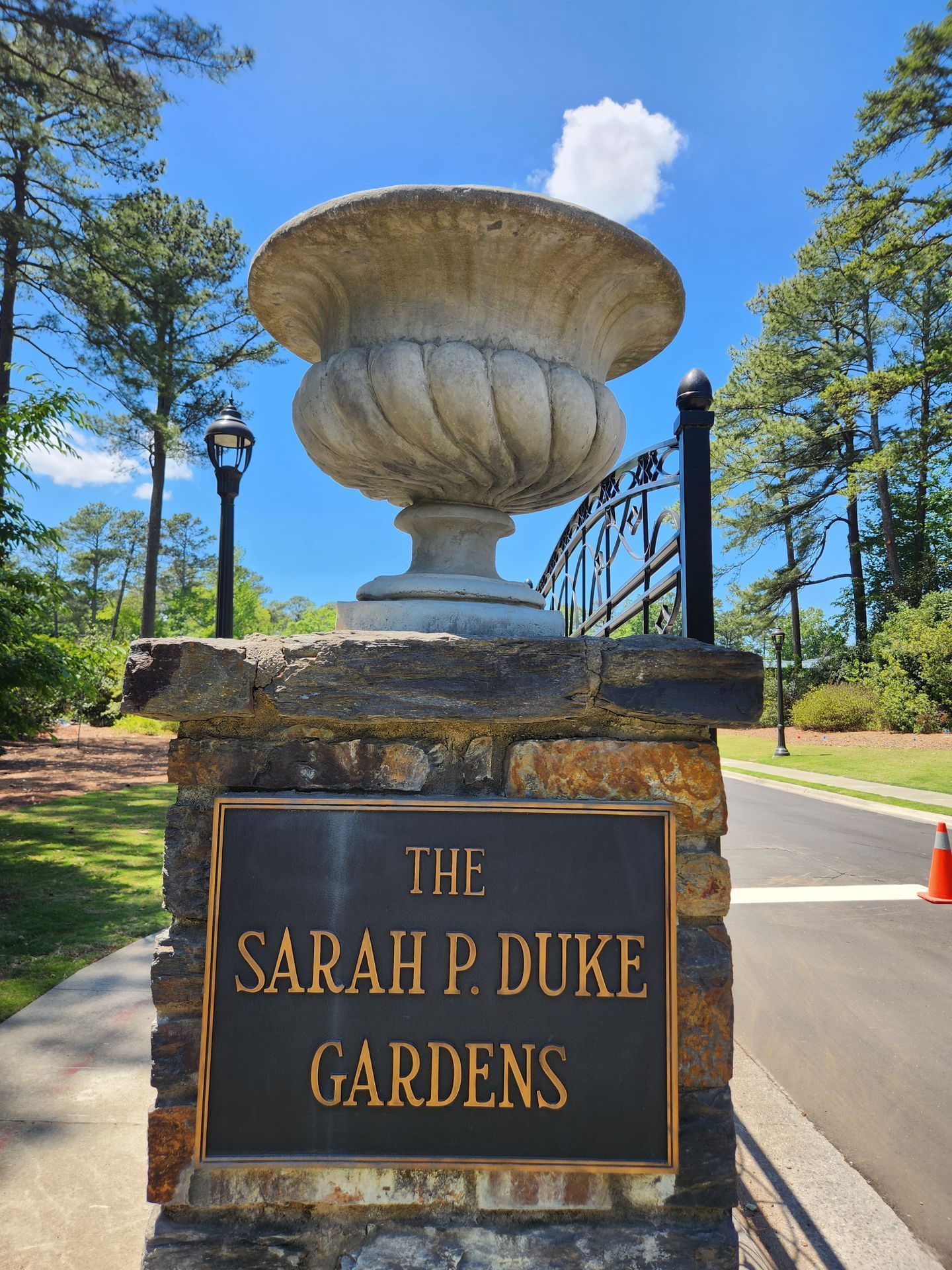 Stone sign for The Sarah P. Duke Gardens beneath a large urn fountain, with trees and blue sky behind.