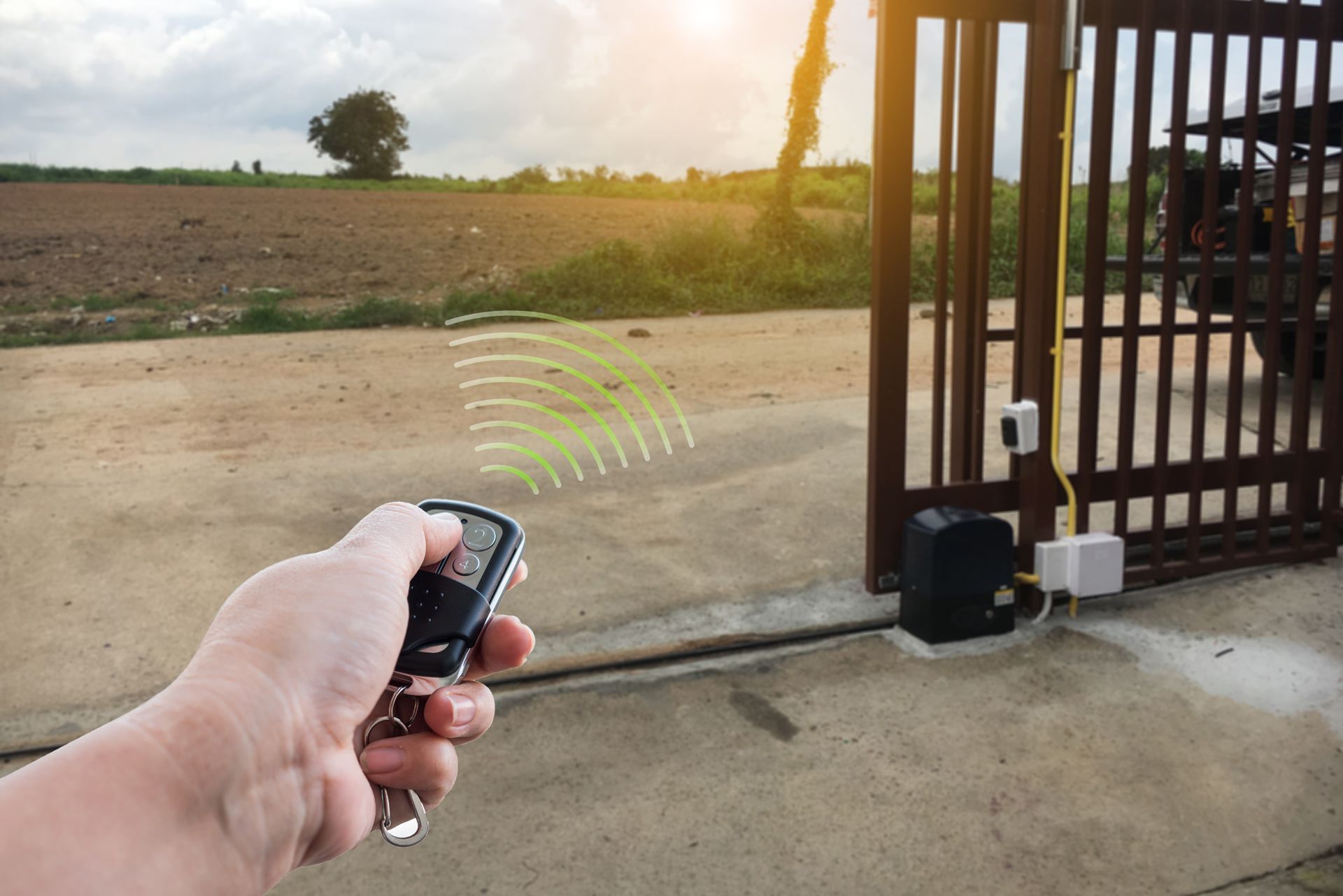 Hand holding a remote pointed at a black gate beside a dirt road at sunset