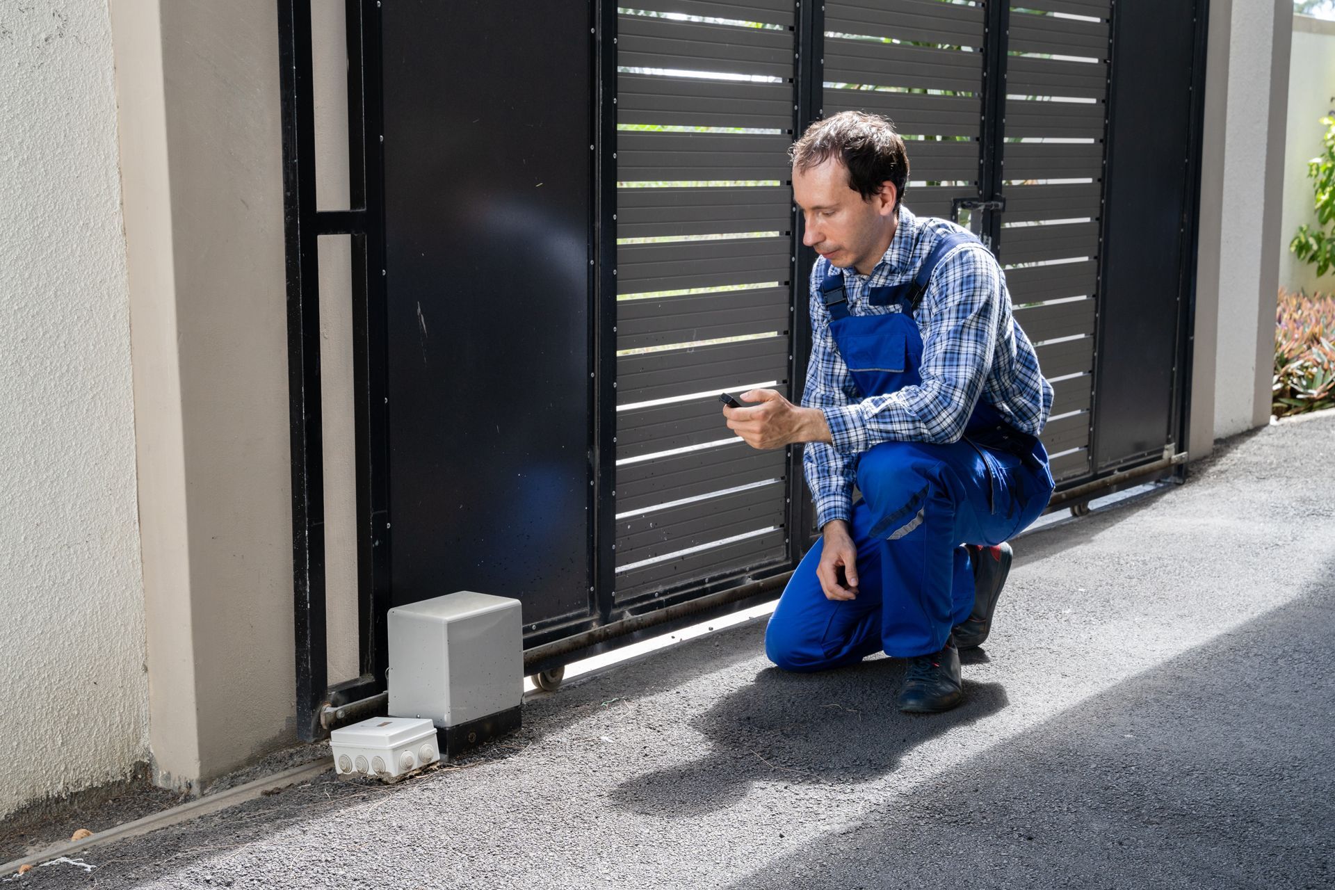 Man kneeling by a black gate, inspecting a small white device on the ground beside a wall.
