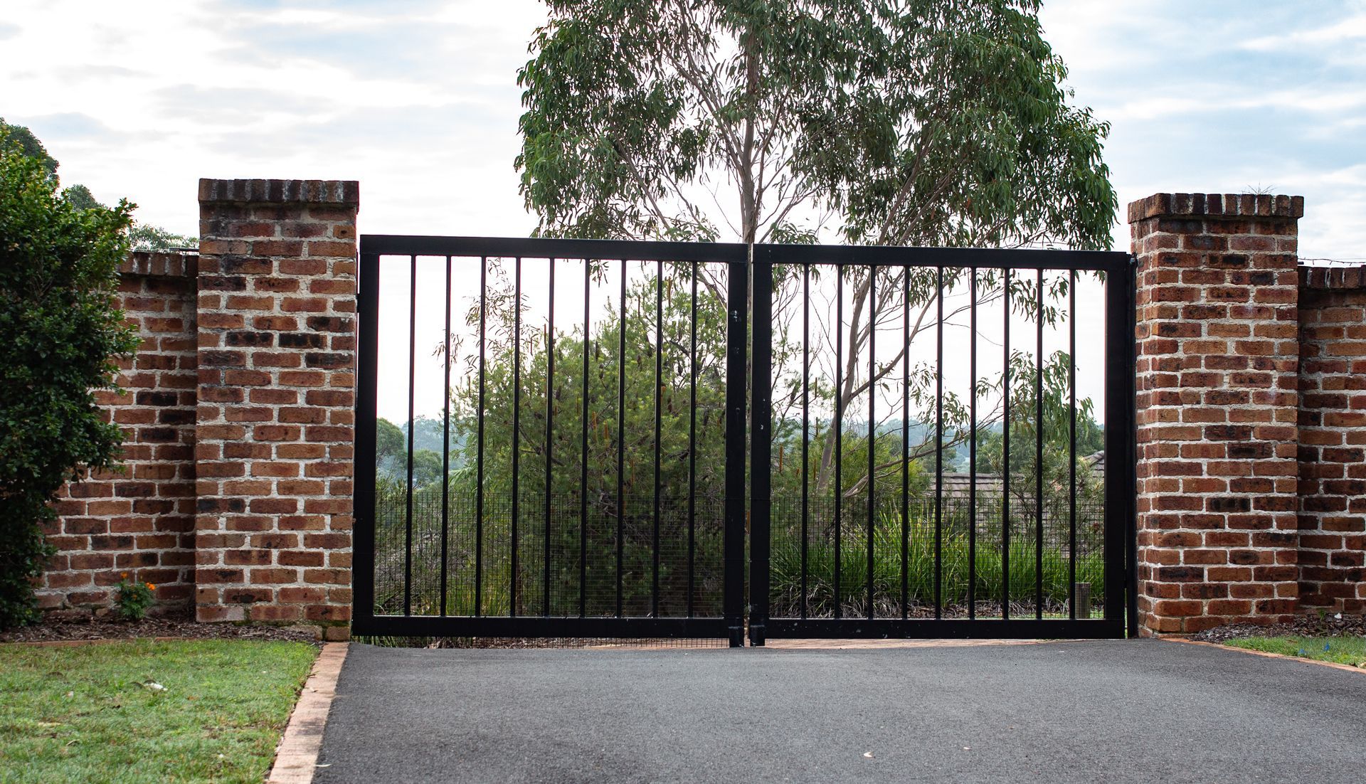 Black metal driveway gate between brick pillars with trees visible behind it