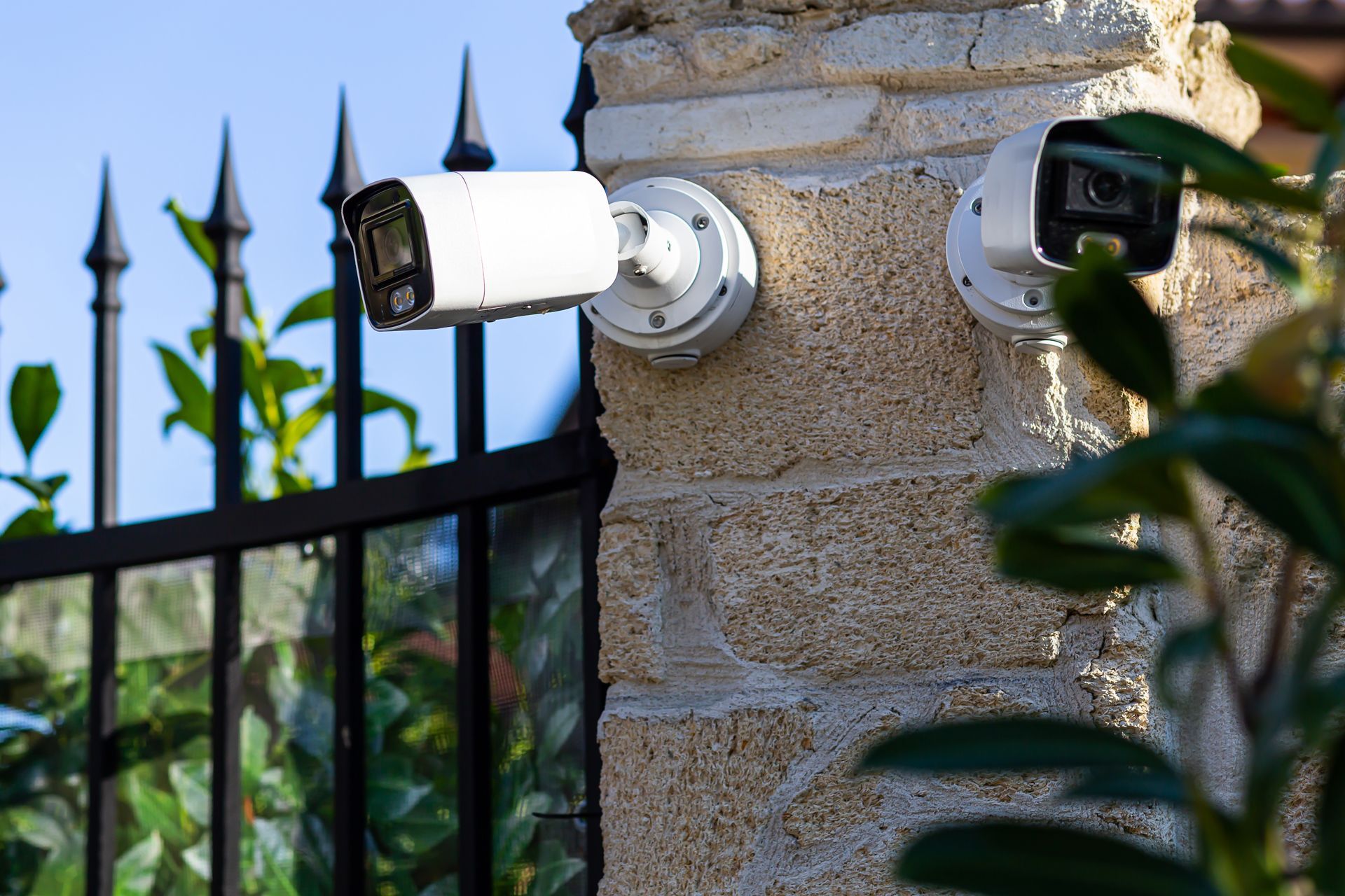 Two security cameras mounted on a stone pillar beside a black iron fence outdoors