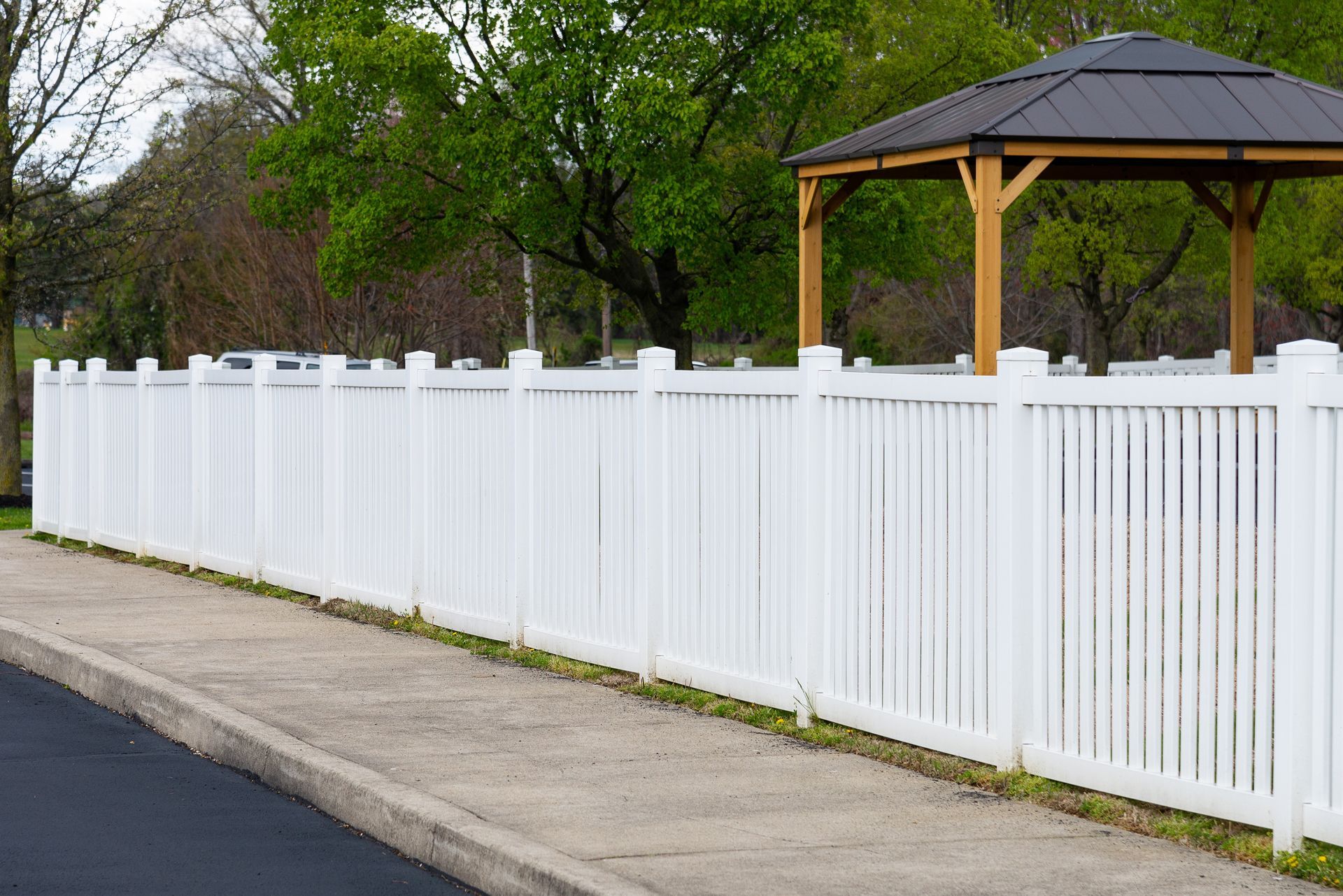 White picket fence along a sidewalk, with a gazebo and trees behind it.
