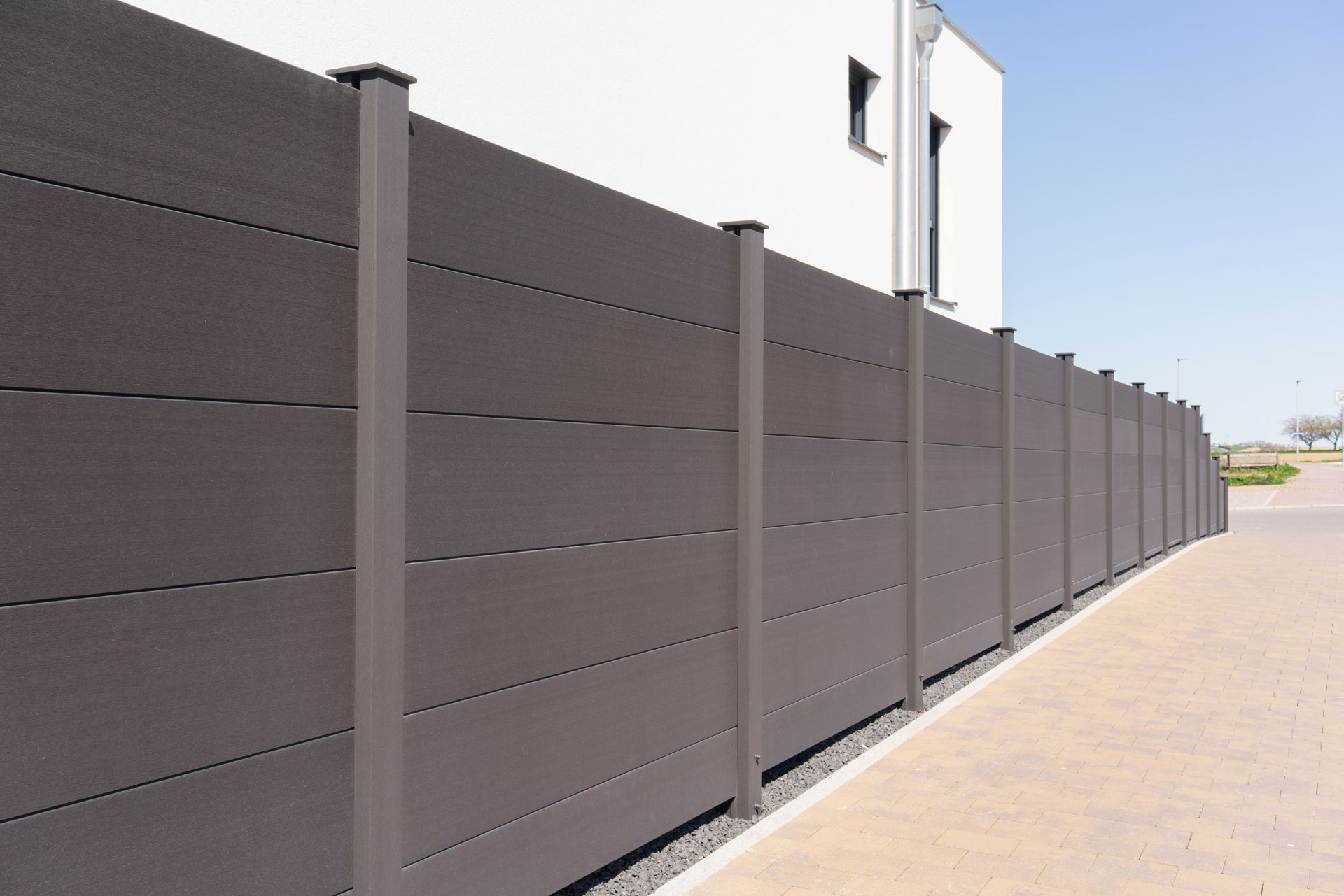Gray privacy fence along a sunlit sidewalk beside a white building