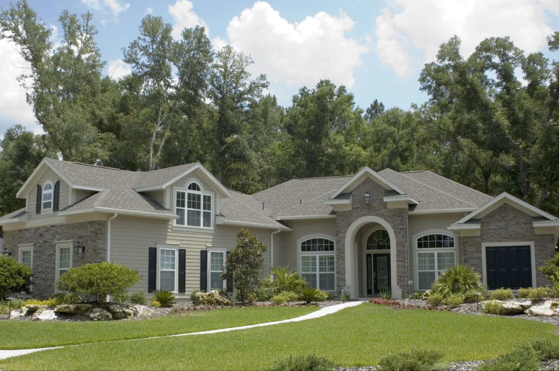 A two-story beige house with gray stone accents, black shutters, and a well-manicured green lawn.