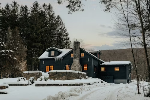 A dark blue house in a snowy forest. Stone chimney, icicles, and a snow-covered driveway.