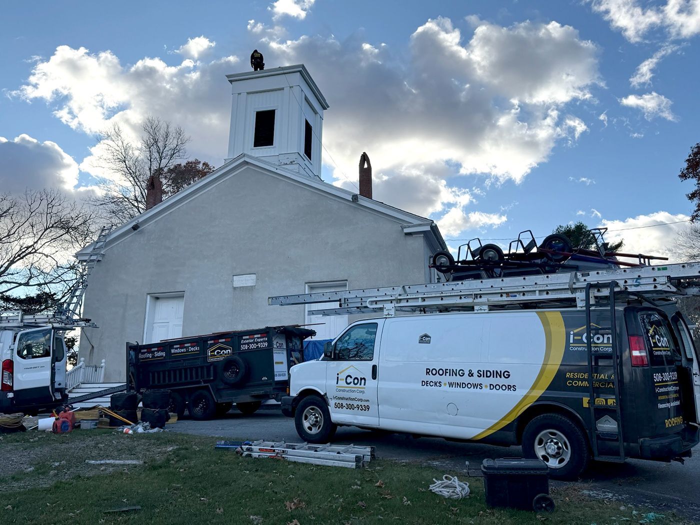 White church with a square tower. Work vans and a dump truck parked nearby; cloudy sky overhead.