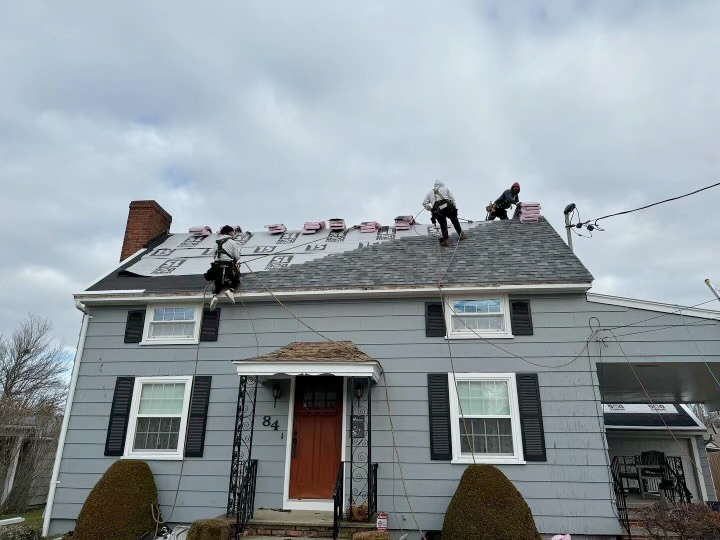 Four roofers working on a gray house roof, under a cloudy sky.